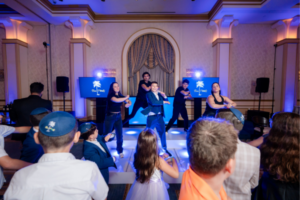 Bat mitzvah boy dancing on stage with a live DJ and professional dancers during a high-energy celebration.