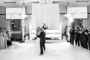 Bride and groom sharing their first dance surrounded by fog and sparkler effects while guests look on.