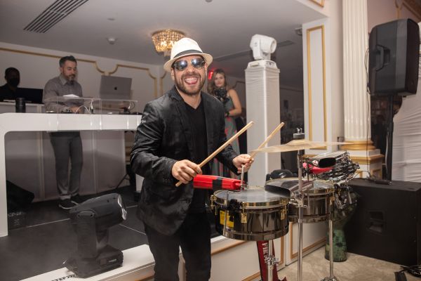 A smiling percussionist in a stylish black outfit and hat playing drums at a wedding, with a DJ and elegant decor in the background.