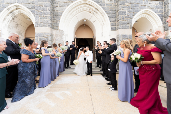 A couple kisses outside a wedding chapel as guests cheer them on.