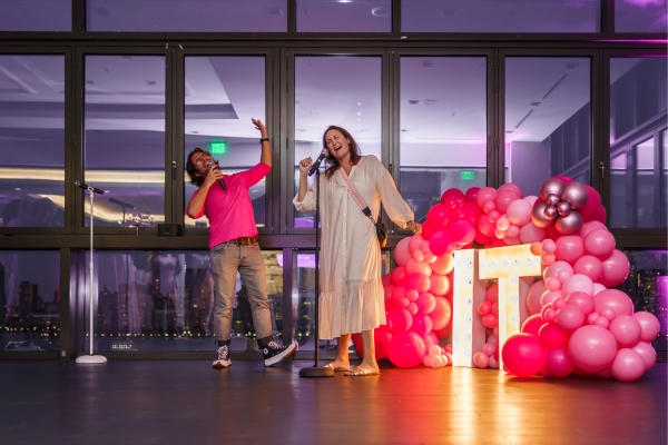Two guests singing a duet on stage with pink balloon decor and a light-up sign.